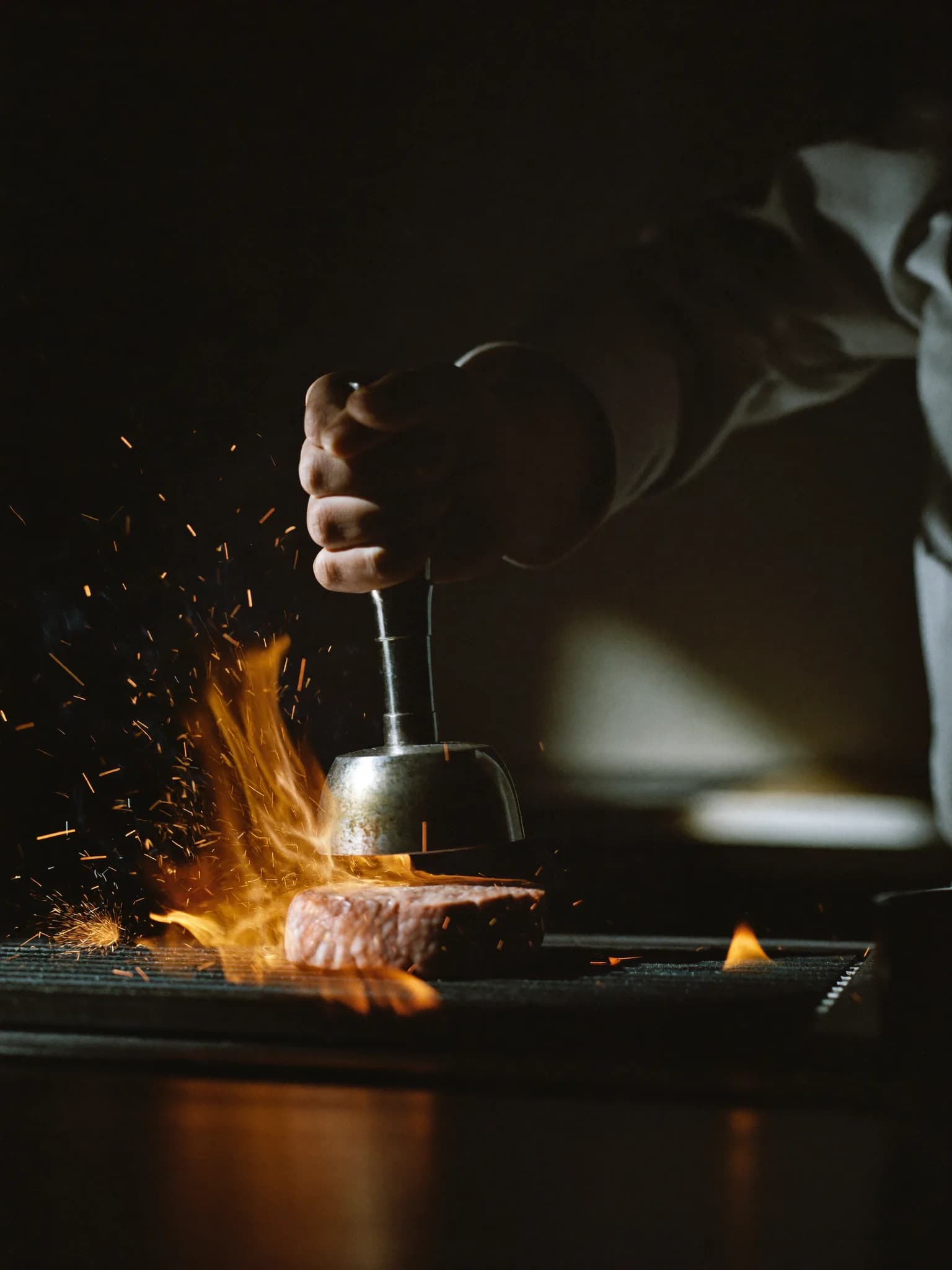 Chef pressing a beef patty onto a flaming charcoal grill, sparks rising
