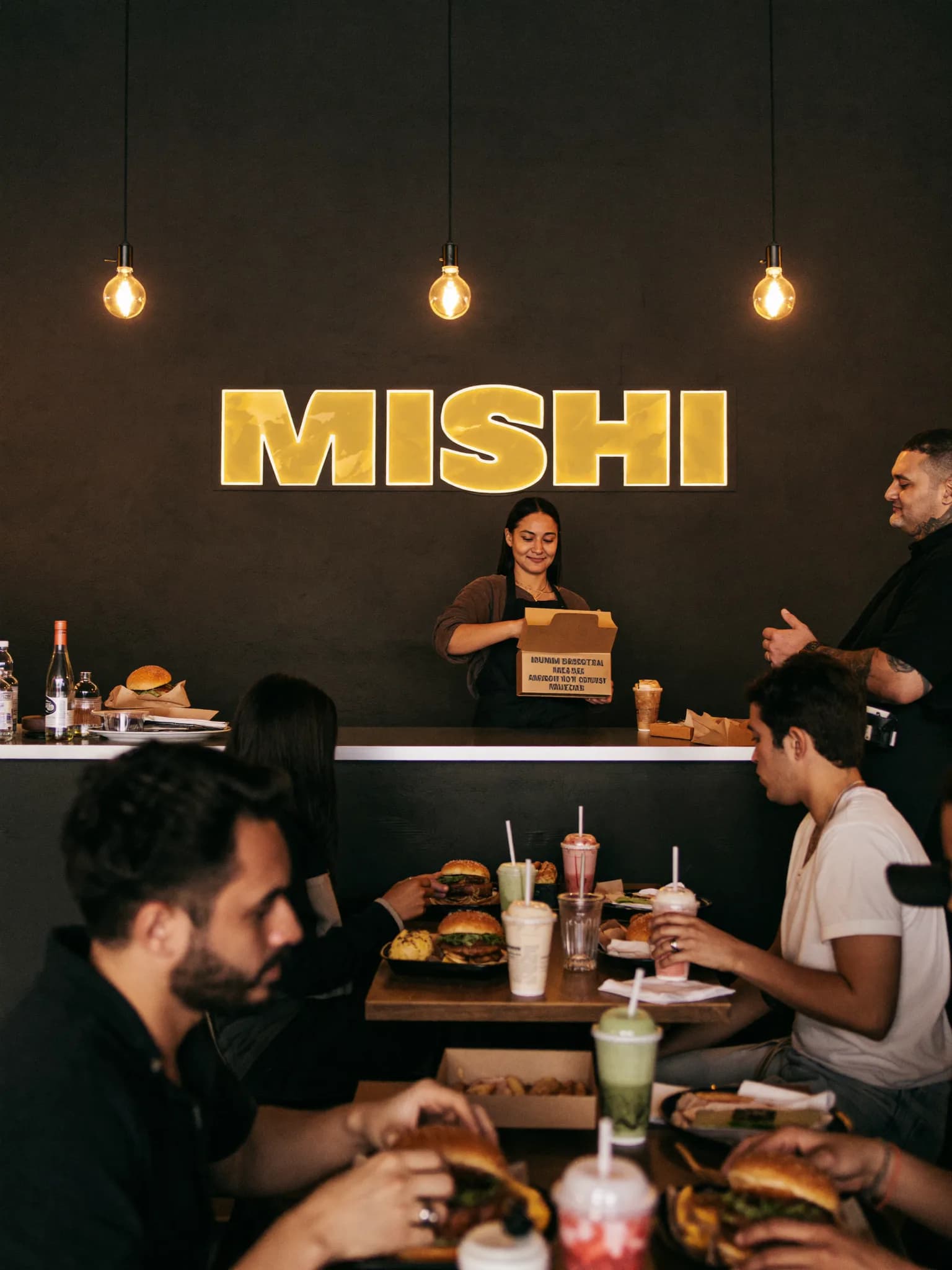 Busy Mishi restaurant with the glowing MISHI sign, customers eating at tables and a staff member serving at the counter